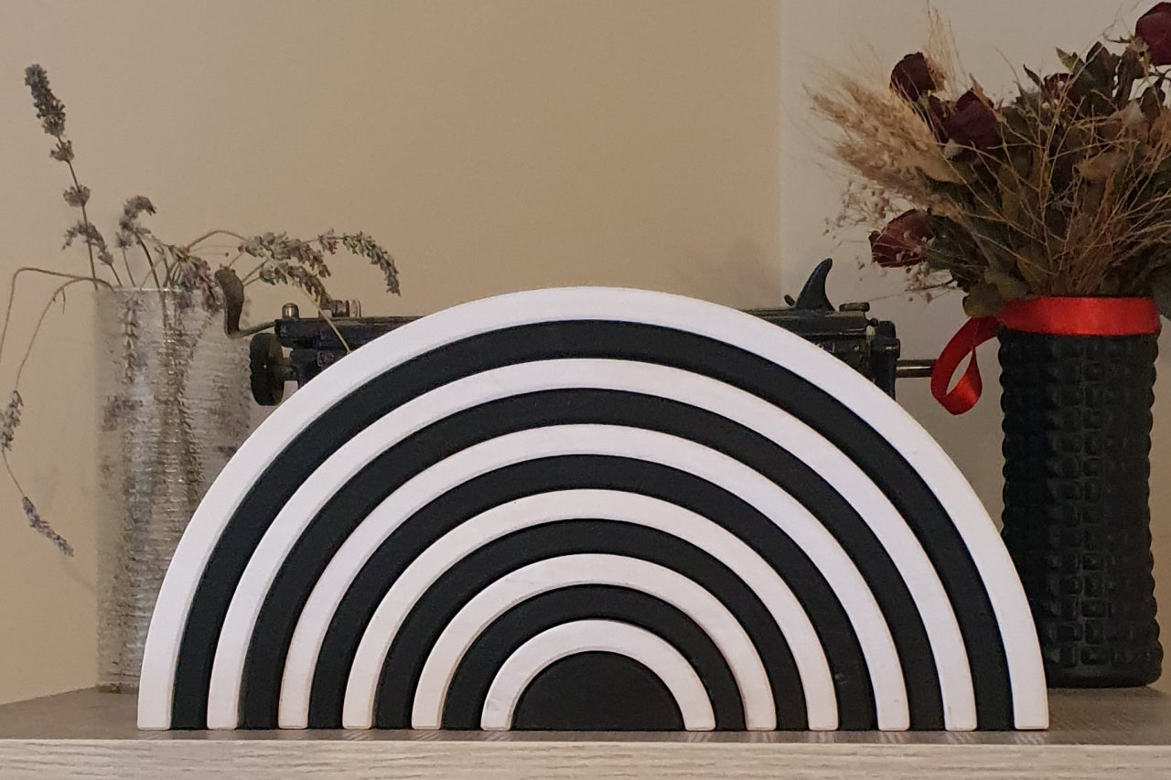 Black and white Waldorf rainbow stacker toy displayed on a shelf with decorative dried flowers and a typewriter in the background.