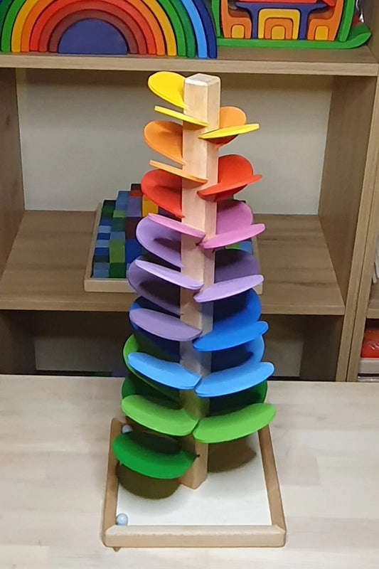 A colorful wooden Waldorf-style musical tree with spiral-shaped leaves in rainbow hues, standing on a wooden base with a marble at the bottom.