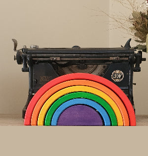 A six-piece colorful Waldorf rainbow wooden arch stacker displayed in front of a vintage typewriter with decorative dried flowers.