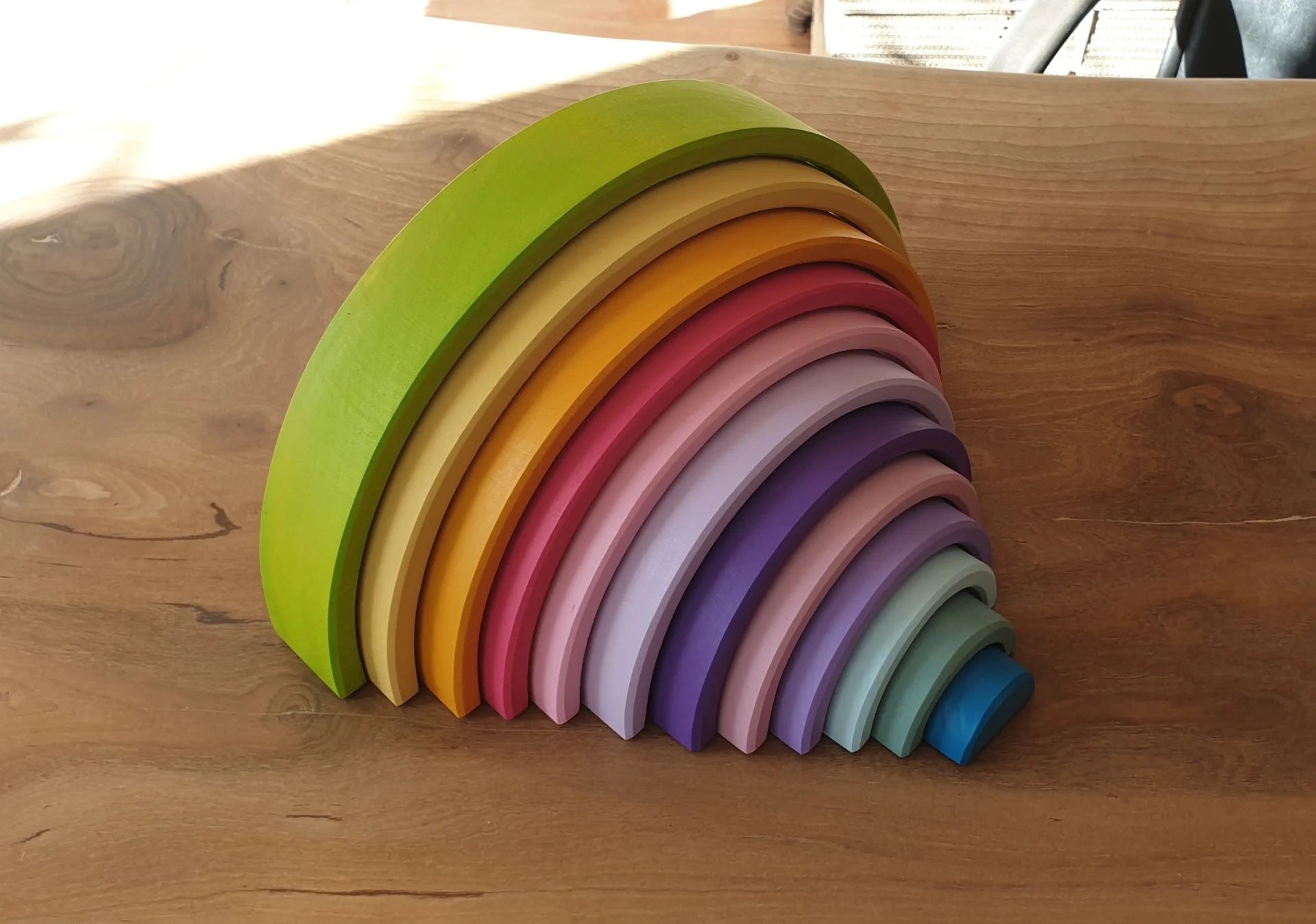 Waldorf rainbow stacker toy arranged neatly on a wooden table under bright sunlight, highlighting its vibrant colors.